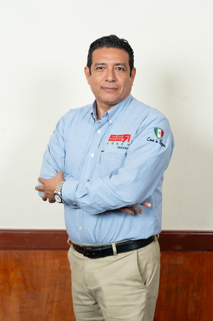 Persona con brazos cruzados vistiendo camisa azul con el logo SER COACHING NEGOCIOS, junto a la bandera de México.
